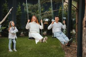 Mom and dad are riding on a swing and there is a daughter standing next to them. The family is resting on a swing photo