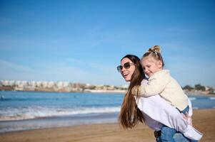 joven amoroso madre dando a cuestas a su encantador niño chica, gasto contento momentos juntos en el playa foto