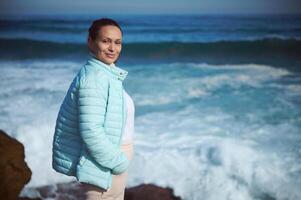 Happy young woman smiles at camera, standing on the edge of a cliff, waves making white foam while breaking on headland photo