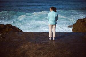 joven mujer disfruta conexión con naturaleza, en pie en rocoso acantilado, contemplando olas salpicaduras rotura en promontorio. foto