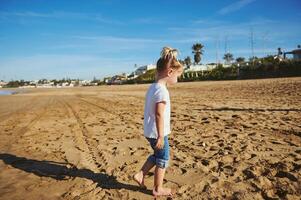 Adorable Caucasian blonde child girl in white t shirt and blue jeans, walking barefoot on the wet sand photo