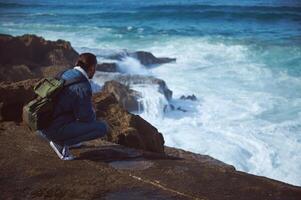 Absorbed male traveler with backpack, admiring the dangerous splashing waves from a rocky cliff photo