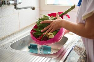 de cerca de hembra manos Lavado alcachofa y verduras debajo fluido agua, en pie por un inoxidable lavabo en hogar cocina foto