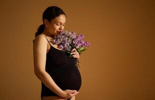 hermosa embarazada mujer en negro mono, participación su grande barriga mientras posando con un ramo de flores de lilas beige antecedentes foto