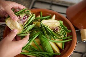 Closeup view of woman's hands putting halves of artichoke flower on a clay dish with vegetables, cooking tagine at home photo