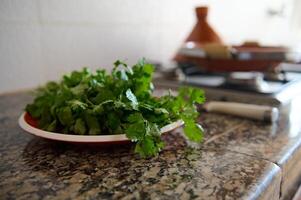 Closeup of coriander and parsley on a plate on marble kitchen counter against the background of tagine cooking on stove photo