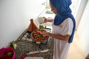Muslim Arab woman housewife with head covered in headscarf blue hijab, stacking fresh vegetables in a dishware from clay photo
