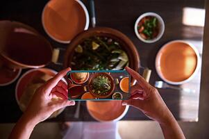 Directly above food blogger hands using smart mobile phone, taking photo of steamed veggies in clay dish tagine