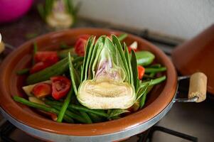 A half of ripe organic green artichoke and vegetables on the clay dish while cooking traditional Moroccan tagine photo