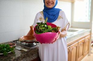 Details on a pink bowl with washed fresh organic vegetables in the hands of a housewife in hijab and authentic dress photo