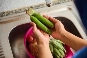 Directly above. Closeup hands holding fresh organic zucchini and washing under running water in the sink at home kitchen photo