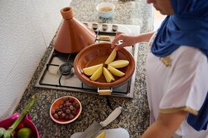 parte superior ver musulmán mujer en hiyab, en pie por estufa en cocina mostrador y apilado patata rebanadas en un tagine arcilla maceta foto