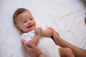 Adorable little baby boy in white bodysuit lying on the bed and connecting with his mother, smiling looking at her photo