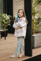 un pequeño niña en un chaqueta con largo pelo es en pie en el terraza a el casa foto
