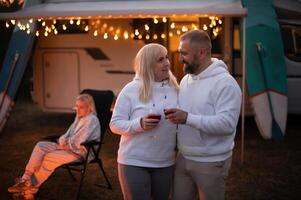 A married couple with glasses of wine stands against the background of a motorhome and rests together by the campfire. Evening family vacation photo