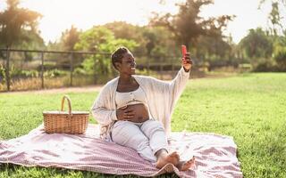 African pregnant woman taking selfie with mobile smartphone while doing a picnic in park - Maternity lifestyle concept photo