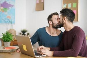 Happy gay couple kissing while using laptop in living room at home - LGBT love and technology concept photo
