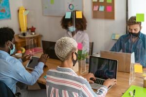 Young multiracial people using computer in co-working creative space while wearing face mask - Health and business technology concept photo