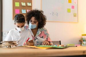 Female teacher with child drawing in preschool classroom while wearing face protective mask during corona virus pandemic - Healthcare and education concept photo