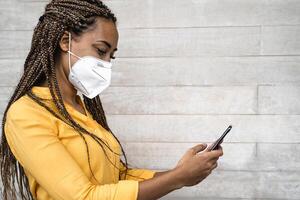 African woman wearing face medical mask using mobile smartphone - Young girl with braids having fun with phone during corona virus outbreak - Health care people and technology concept photo