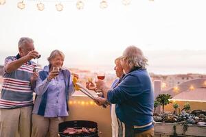 Happy senior friends having doing barbecue party on patio - Retired people having fun drinking wine and grilling meat at bbq dinner at home - Friendship, food and elderly lifestyle concept photo