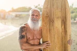 Senior male having fun surfing during sunset time - Fit retired man training with surfboard on the beach - Elderly healthy people lifestyle and extreme sport concept photo
