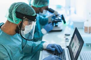 Doctors wearing personal protective equipment working in laboratory using computer and examining coronavirus through microscope - Science and technology concept photo