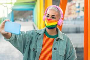 Young woman wearing gay pride mask listening to music with wireless headphones and taking selfie with mobile smartphone outdoor - Gender equality and technology concept photo