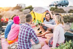 Group of friends cheering with beers at barbecue dinner on the beach - Happy people camping with tent and having fun toasting bottles of beer at bbq - Friendship and youth lifestyle vacation concept photo