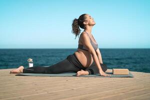 Young pregnant woman doing prenatal pilates exercises session next the sea - Health lifestyle and maternity concept photo