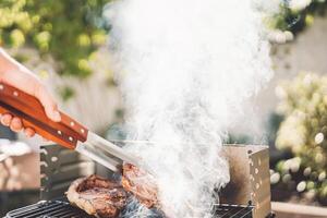 Man chef grilling beef steak at barbecue dinner party outdoor - Close up male hand cooking meat on bbq for family in the garden - Food, weekend activities and people lifestyle concept photo