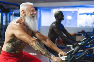 Senior and young men doing bike exercises in gym - People with different age and race training on cycling machine in health wellness center - Sport activity and fitness lifestyle concept photo