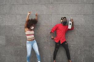 joven africano personas vistiendo cara máscara y bailando al aire libre mientras escuchando a música con inalámbrico auriculares y Clásico boombox foto