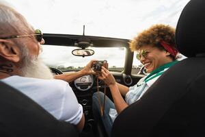 Happy senior couple having fun taking photo with old vintage camera while driving on new convertible car during road trip tour vacation