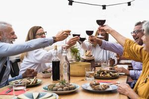 multirracial mayor amigos teniendo divertido comida juntos y tostado con rojo vino en casa patio cena - comida y Días festivos concepto foto