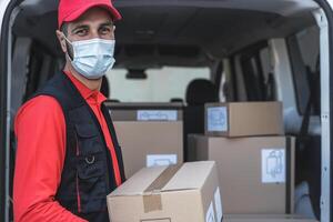 Young delivery man carrying cardboard box while wearing face mask to avoid corona virus spread - People working with fast deliver during corona virus outbreak photo