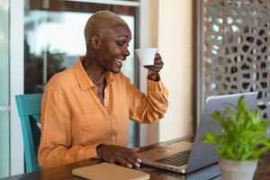 Afro senior woman having fun doing video call using computer while drinking coffee in bar restaurant photo