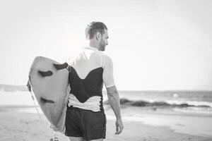 Young surfer holding his surfboard on the beach - Handsome man waiting waves for surfing - Black and white editing - people, sport and lifestyle concept photo