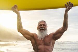 Happy fit senior having fun surfing at sunset time - Sporty bearded man training with surfboard on the beach - Elderly healthy people lifestyle and extreme sport concept photo