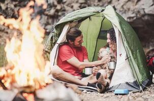 Happy trekker couple camping tent with dog next bonfire  - Hipster man and woman having fun mountaineering together - Love relationship and travel lifestyle concept photo