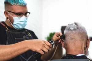 Male hairdresser cutting hair to hipster senior client while wearing face surgical mask - Young hairstylist working in barbershop during corona virus outbreak - Health care and haircut salon concept photo
