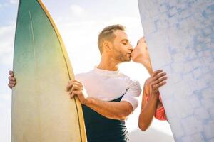 salud joven Pareja de surfistas besos a puesta de sol en el playa participación tablas de surf - contento amantes teniendo un oferta momento con un Beso mientras surf juntos - gente, amar, deporte y estilo de vida concepto foto