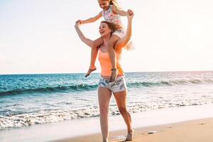 contento amoroso familia madre y hija teniendo divertido en el playa en vacaciones - mamá que lleva en espalda su niño y corriendo siguiente a Oceano - vacaciones, padre, materno amor concepto foto