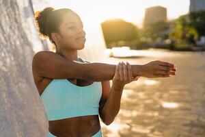 Young African woman doing warming up exercises before workout session in the city at sunset - Sport people lifestyle concept photo