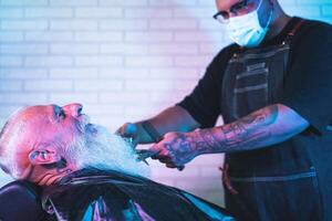 Male hairdresser cutting beard to hipster senior client - Young hairstylist working in barbershop - Haircut profession concept photo