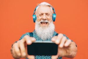 Senior bearded man making selfie with mobile phone while listening to his favorite playlist with headphones - Fashion matured male having fun with smartphone apps outdoor - Elderly, technology concept photo