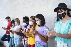 Young people wearing face mask using mobile smartphone outdoor - Multiracial friends having fun with new technology social media app during corona virus outbreak - Youth millennial lifestyle concept photo