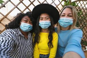 Multiracial friends wearing face mask while taking selfie with mobile smartphone cam during corona virus outbreak - Soft focus on central girl photo