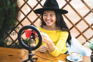 contento asiático mujer vlogging en línea utilizando teléfono inteligente leva y hombre de influencia LED mientras Bebiendo un café sentado en bar foto