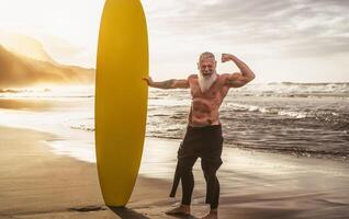 Happy fit senior having fun surfing at sunset time - Sporty bearded man training with surfboard on the beach - Elderly healthy people lifestyle and extreme sport concept photo